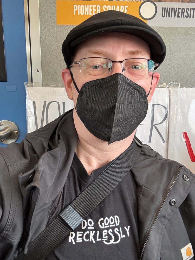 Me, a white man with glasses, wearing a black cap, face mask, and jacket, with a black t-shirt that says “Do Good Recklessly”. 