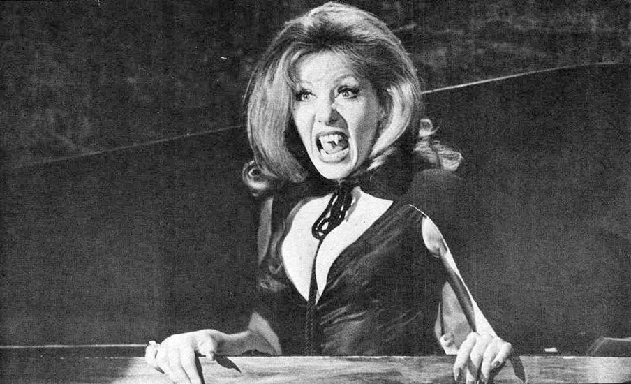 b/w still of Ingrid Pitt in Countess Dracula. She is wearing an extravagantly low-cut dress, and fangs, and is rising from a coffin with her mouth wide open in a predatory if slightly comical manner