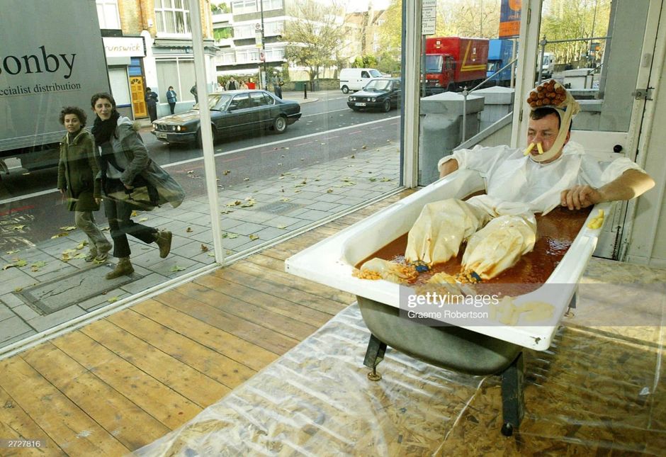 Two people walk past a large plate glass window looking in, possibly scornfully, at a man in white clothes sitting in a bath full of baked beans. He has crinkle cut chips up his nose and a number of sausages strapped in a bundle to the top of his head