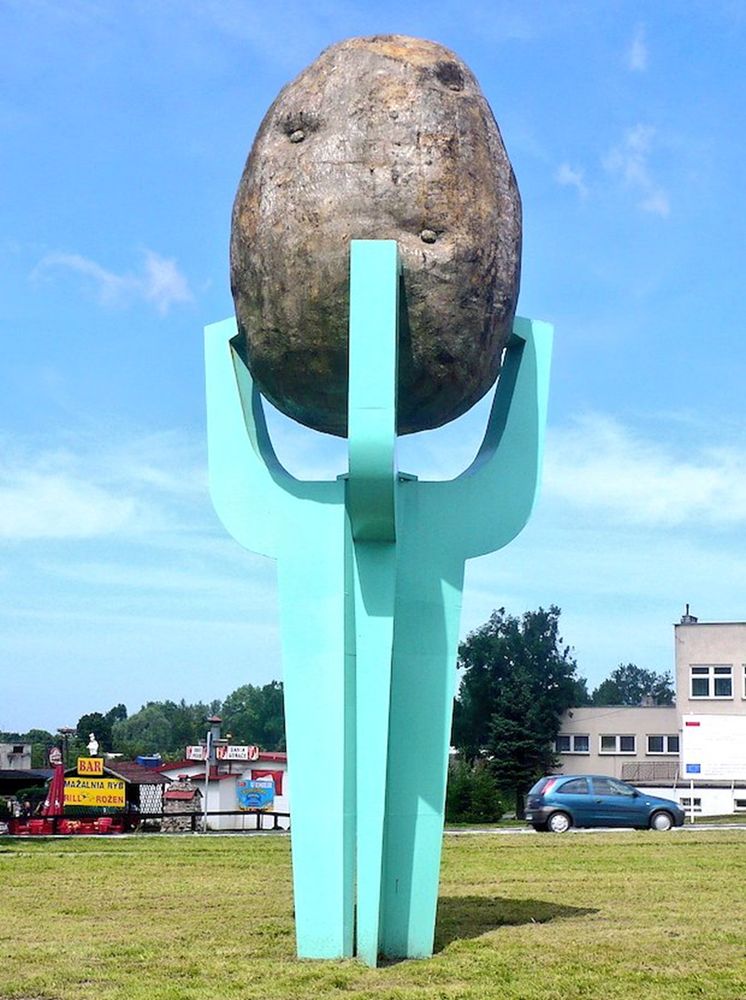 A large sculpture in a park, possibly, or at least an area of grass by a road, in Poland. An enormous pale blue stand, with four prongs, holds up a gigantic (metal?) potato, complete with knobbles