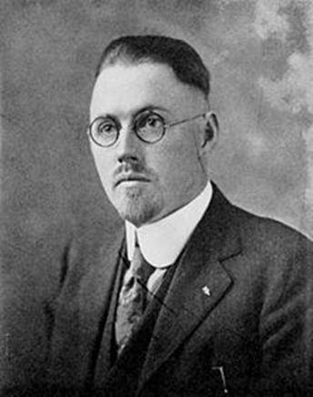 A man with slicked back hair and one of those shaved round the back and sides haircuts. He’s wearing round glasses, and a stiff, rounded collar, and has a goatee. A wrong ’un if ever I saw one