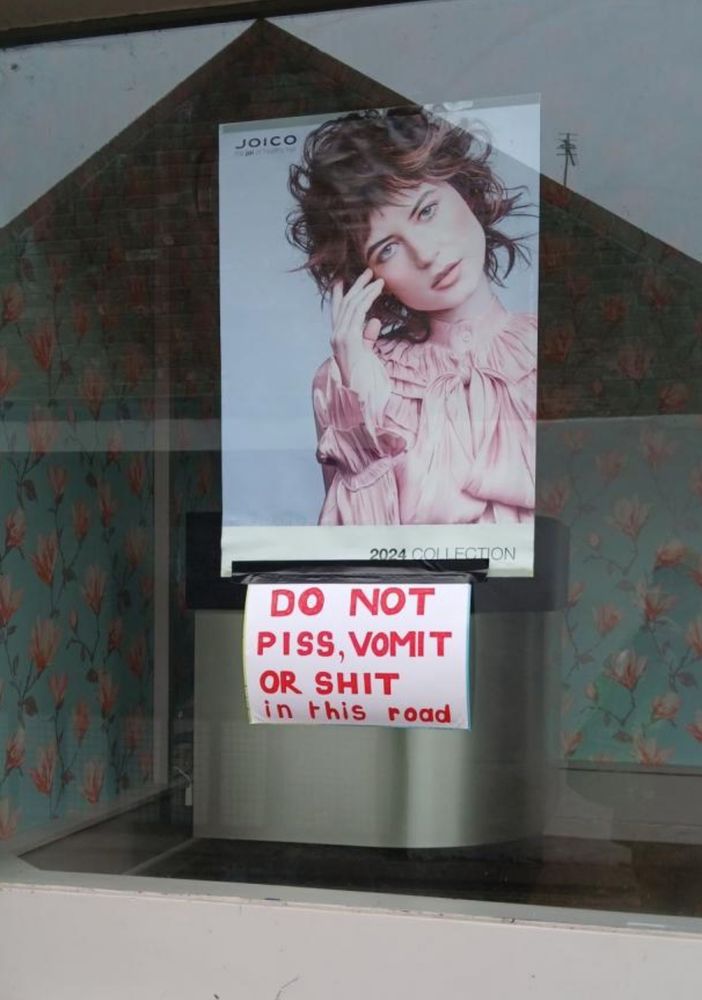 A poster in a disused hairdresser’s shop showing a woman modelling for a hair product. Underneath, a handmade sign has been fixed to the poster with black tape. It says “DO NOT PISS, VOMIT OR SHIT in this road”