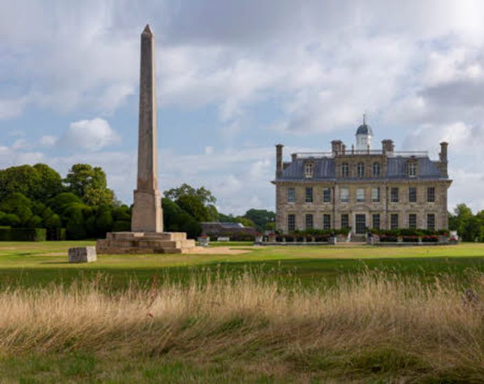 An obelisk in the grounds of a country house, with grass in the foreground, and trees and a cloudy sky behind