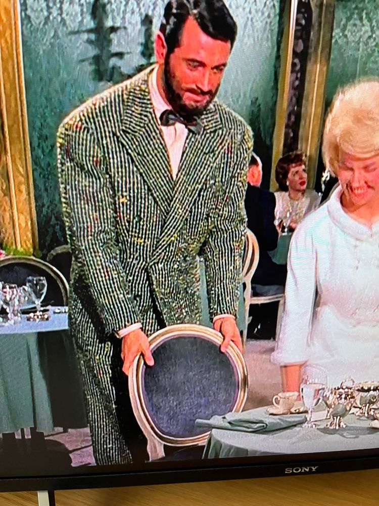 Rock Hudson moving a chair for Doris Day in Lover Come Back. He has a beard, and is wearing an extraordinary green suit with stripes and flecks of red, blue, and yellow in it