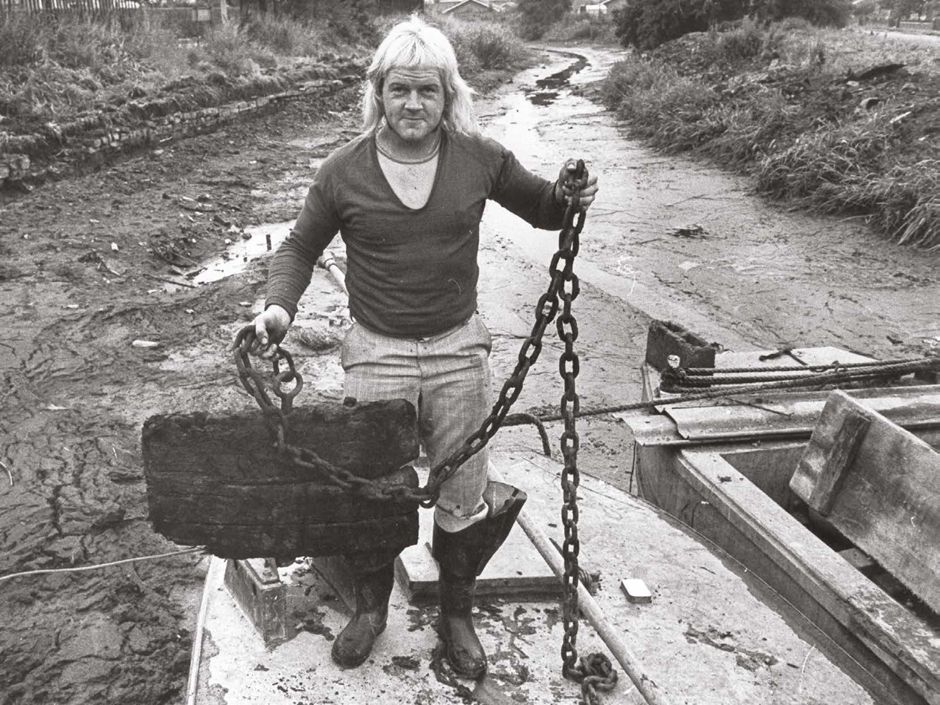 A man with a spectacular 1970s mullet, wearing big wellies and a v-neck jumper over a T-shirt, looks at camera as he stands on a boat which is itself standing on the muddy bottom of an empty canal. He is holding up an enormous wooden plug on a long chunky chain