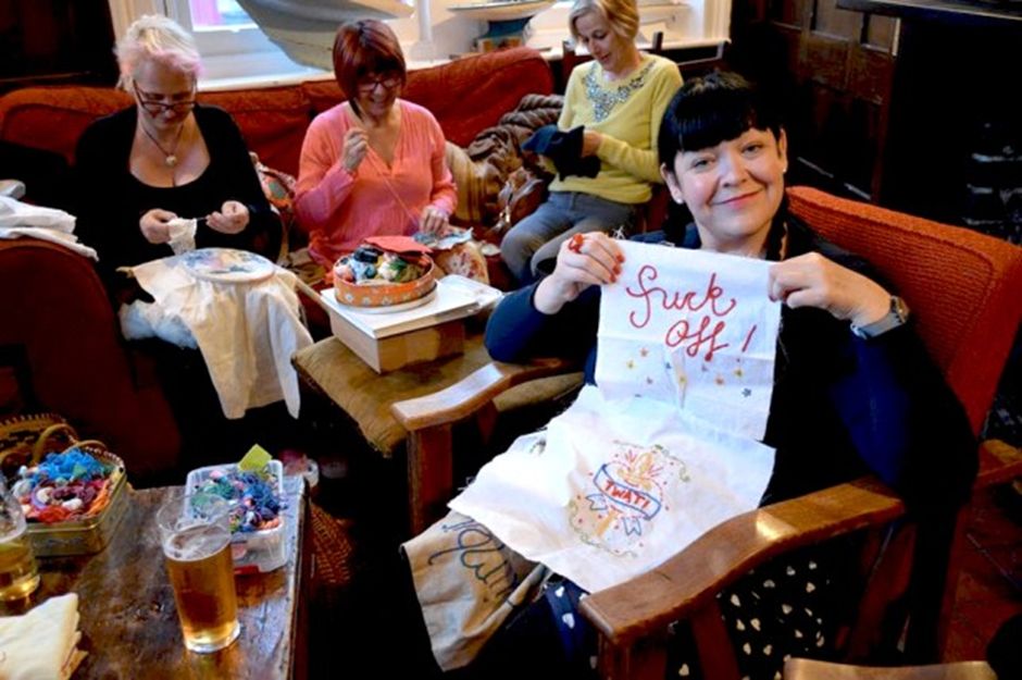 Members of the Whitstable’s Profanity Embroidery Group at the Duke of Cumberland pub. Three women sit on a sofa, sewing, while a woman in front, with a pint on the table in front of her, holds up a piece of fabric on which she has stitched the phrase ‘fuck off!’ Below that, another design reads ‘TWAT!’ She is smiling proudly