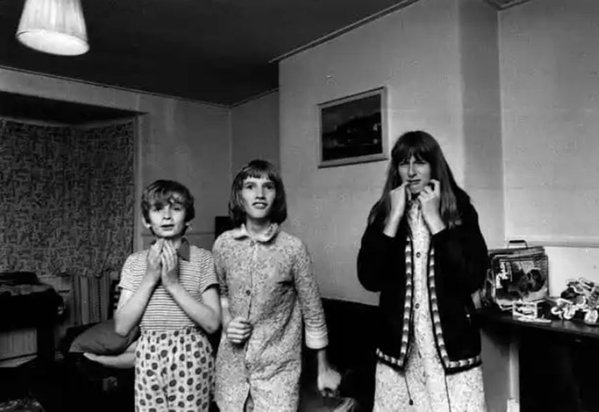 b/w photo of three children in nightclothes. A young boy, left, looks frightened, with his hands clasped together under his chin. An older girl on the right has her hands up at her face, with her little finger in the corner of her mouth. Janet, in the middle, looks excited
