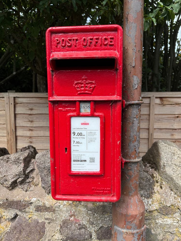 A red post box attached to a lamppost with peeling paint with a wall and fence behind.
