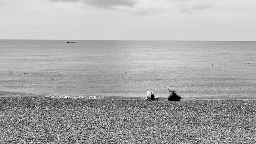 Monochrome photograph of a shingle beach with two people lying on it with legs crossed and a boat offshore in a clam sea. 