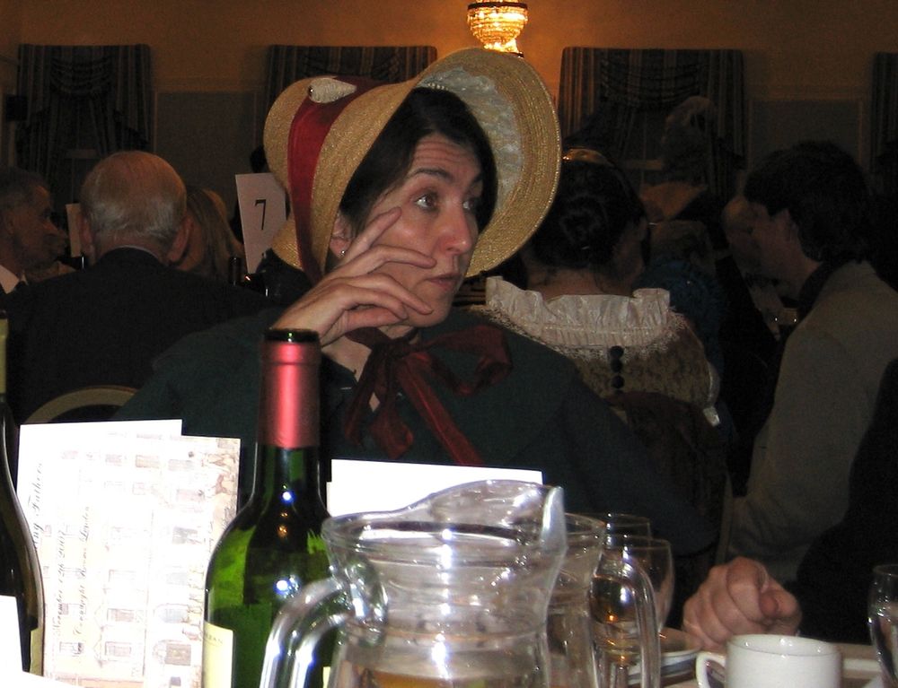 Photograph taken across a dinner table with glasses and a wine bottle, a woman wears a straw bonnet tied with a red ribbon, and a green dress. 