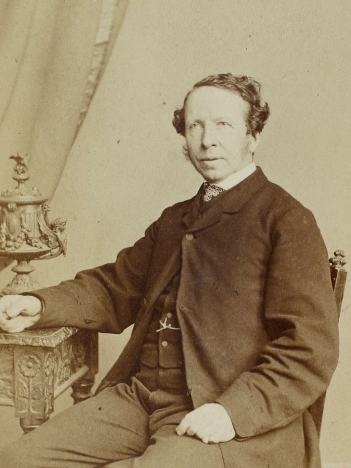 Photograph of Thomas Hawkins sitting at a table and wearing a dark jacket and waistcoat.