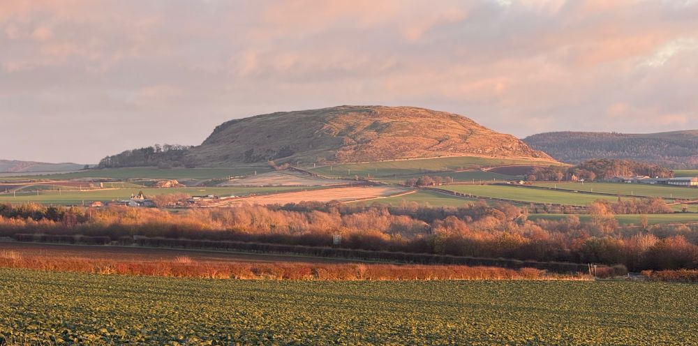 View across fields and trees to a rounded hill formed of an early Carboniferous laccolith, a domed igneous intrusion, lit by today's warm afternoon sunlight.