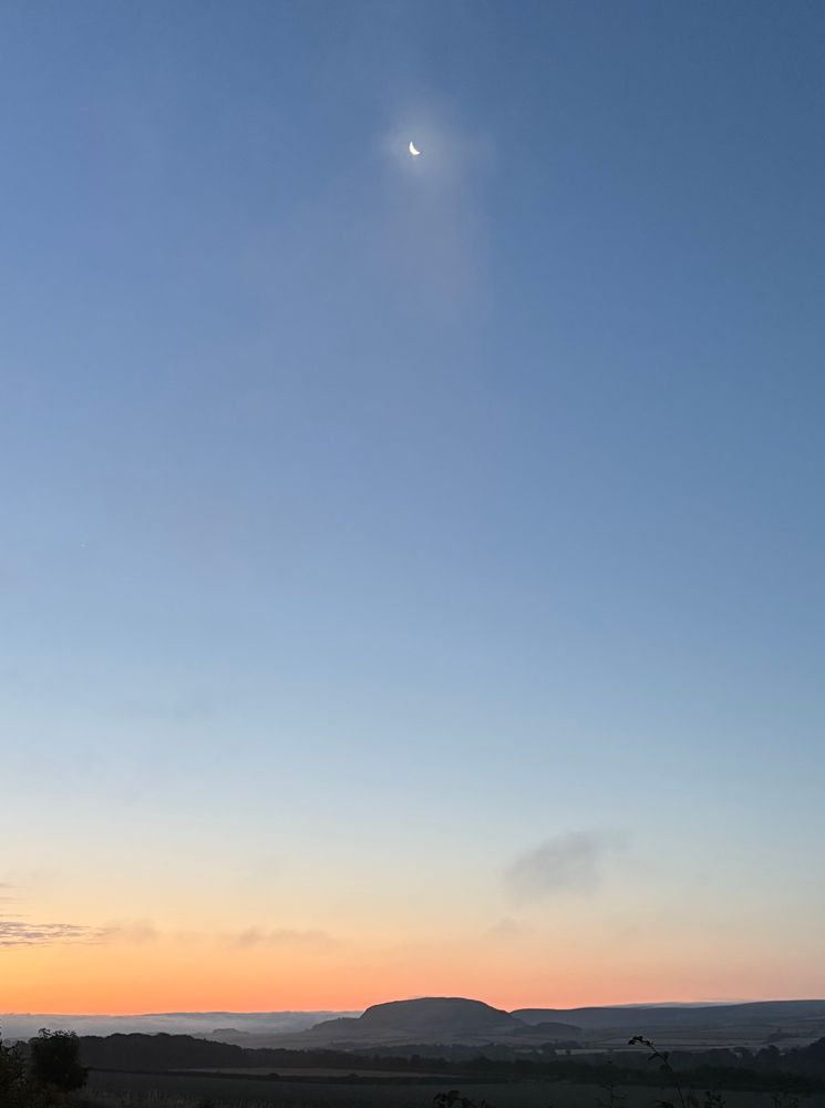 A crescent moon before dawn over a round hill 