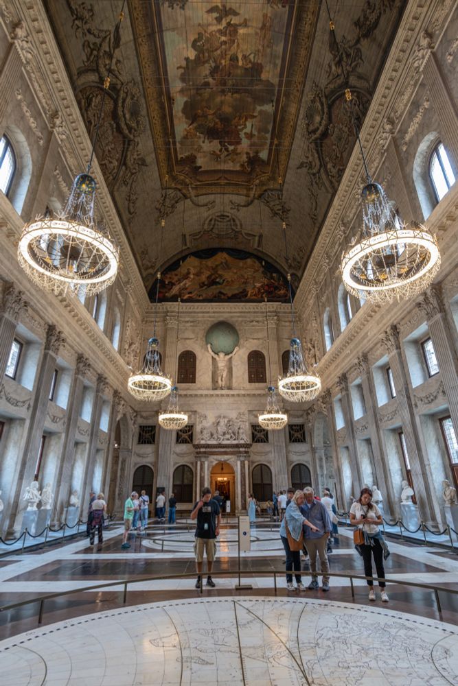 A photograph of the Citizen's Hall in the Paleis Amsterdam. A few people in the foreground are looking at one of the maps inlaid into the floor; six chandeliers hang three to each side below the ceiling, which is painted with several scenes, and Atlas stands over the far end of the hall, holding the Earth on his shoulders. 
