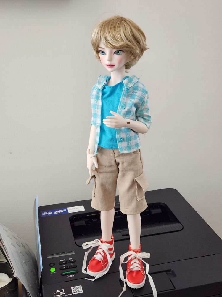A doll with blonde hair and blue eyes wearing a blue t-shirt, blue-and-white short-sleeved button-down, khaki cargo shorts, and red sneakers 