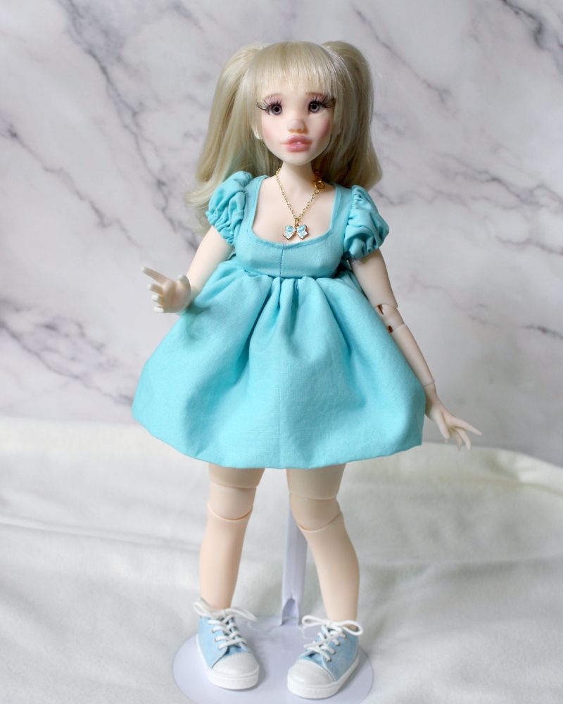 A doll with blonde hair and blue eyes wearing a light aqua dress and blue sneakers