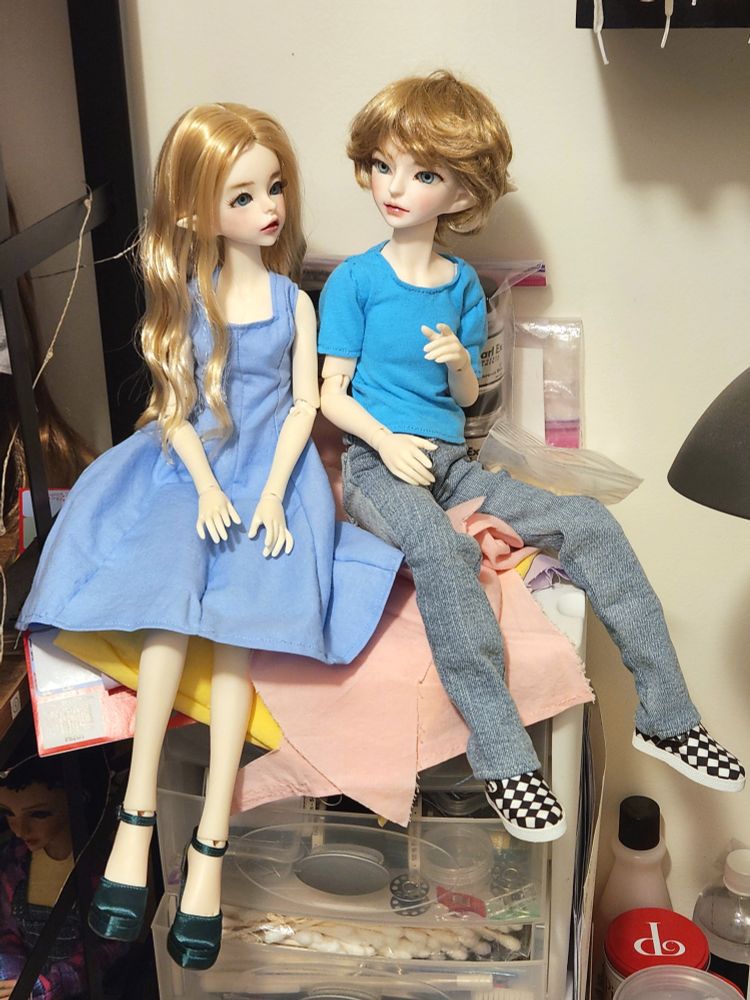 Two dolls with blonde hair and blue eyes sitting on top of a set of drawers. One doll is wearing a blue dress and green heels. The other is in a blue tee shirt, jeans, and checkered Vans.