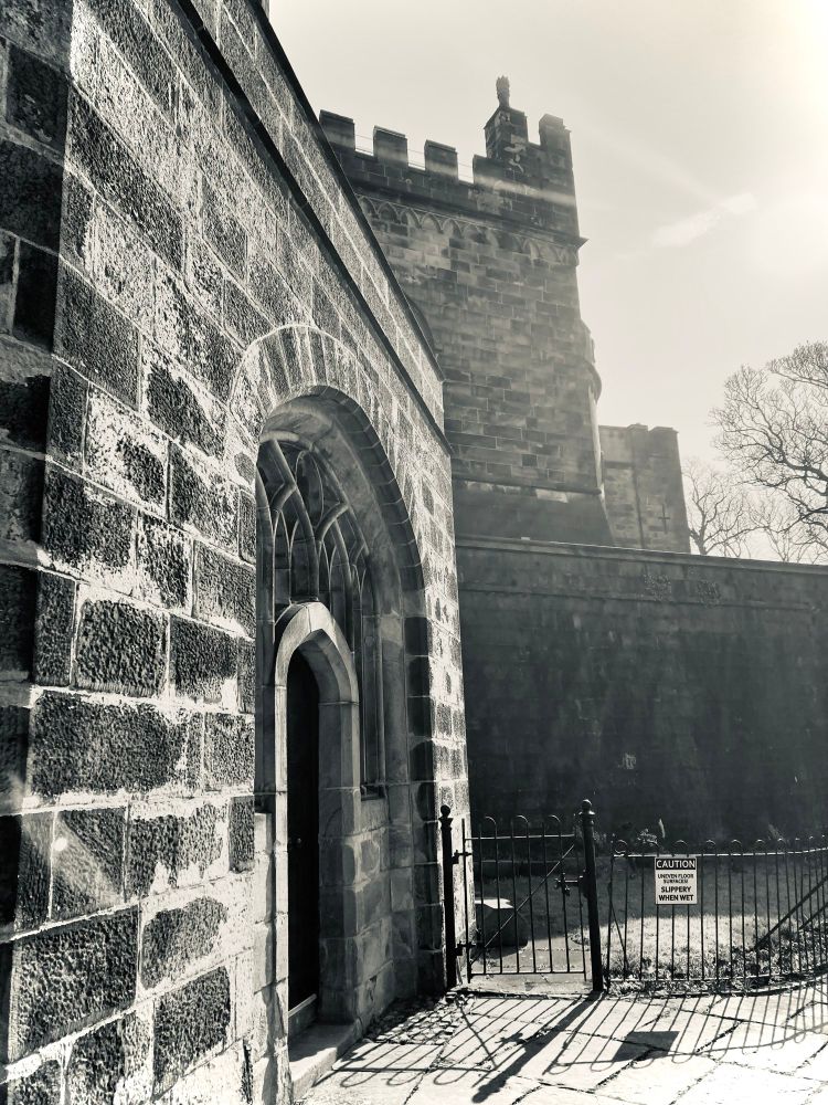 Black and white photo of a small side door with fancy pants arched windows around it, and a slice of castle walls, looking old and broody in angled sunlight
