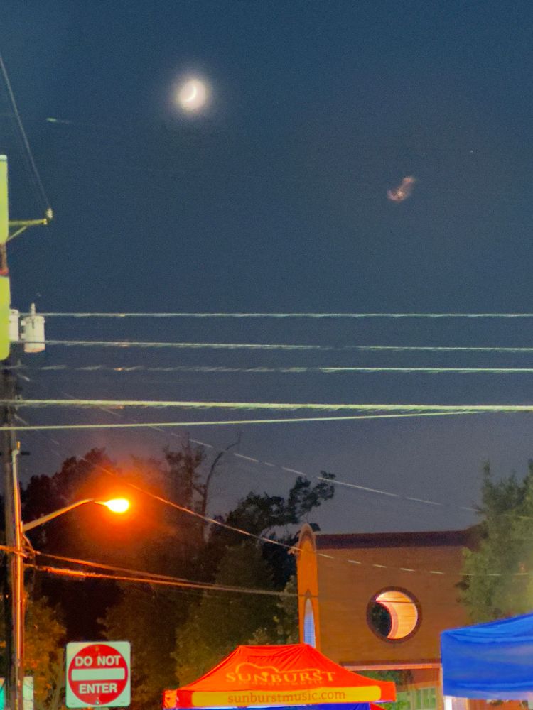 the real moon in a crescent shape in upper left corner of photo and a 2nd story round hallway cutout that looks like a crescent moon from my vantage point in the lower right corner. streetlight and a red and white “do not enter sign in mid right of shot and a red and yellow event tent at the bottom of the shot 