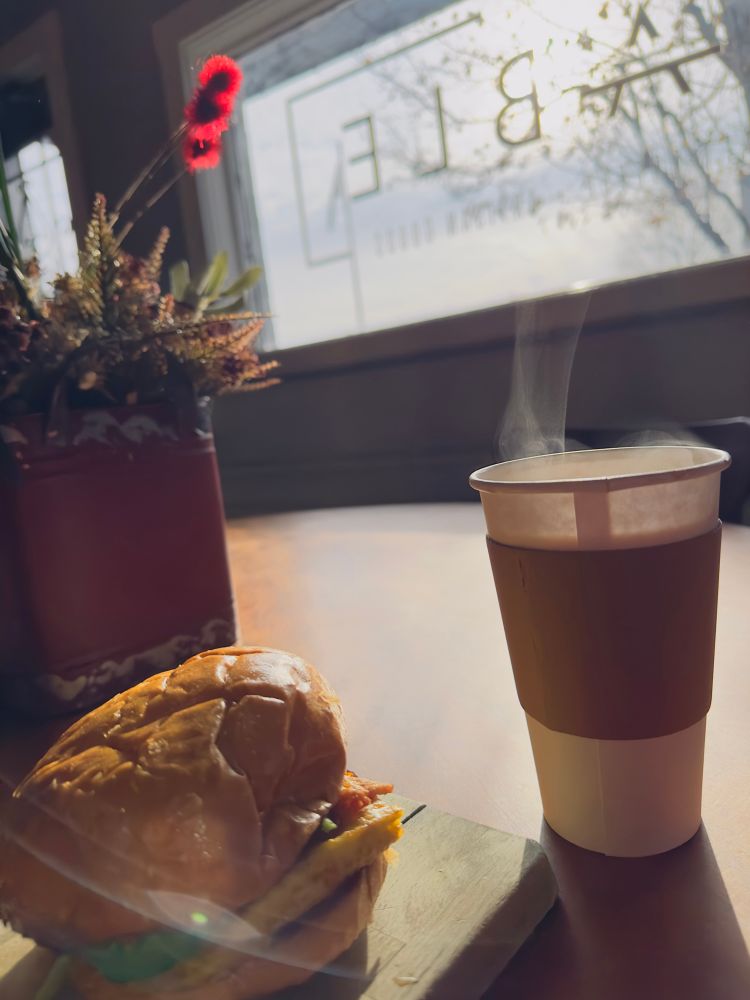 a breakfast sandwich on a mini cutting board and a paper coffee cup with a sleeve throwing off steam on a sunlit table in front of a coffee shop window (“Table” in Washington, PA)