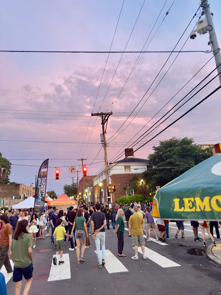 street festival with many tents and stands with food and craft vendors crosswalk in the miscount and many people out enjoying the market. sky is pink and purple 