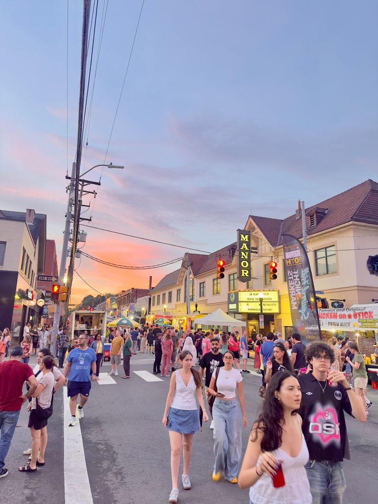 street festival with many tents and stands with food and craft vendors crosswalk in the miscount and many people out enjoying the market. sky is pink, orange, blue, and purple 
