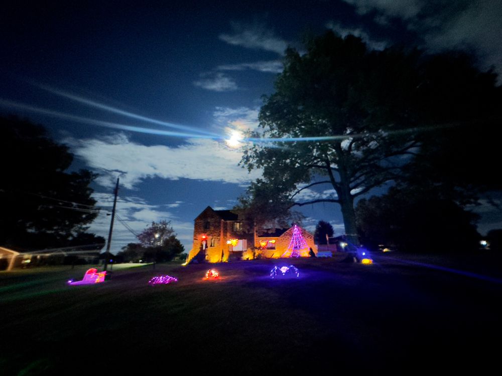 a large stone house with purple, orange, and red illuminated halloween decorations (spider, coffin) in the yard, under a midnight blue sky with a bright white full moon over light blue clouds. a large tree is in silhouette to the right 