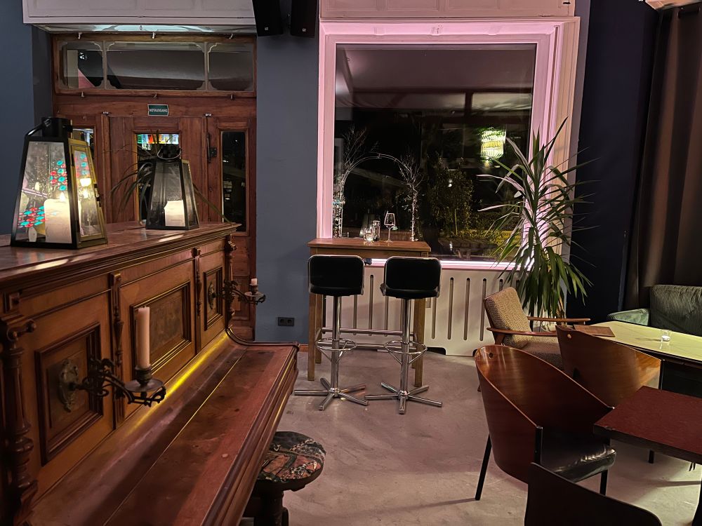 Ein gemütliches Interieur einer Bar oder eines Cafés mit einem Klavier mit Kerzenleuchtern, hohen Hockern und verschiedenen Sitzgelegenheiten. Große Fenster bieten einen Blick auf das Äußere, das durch weiches Licht beleuchtet wird. Eine Pflanze ist in der Ecke sichtbar, was zum Ambiente beiträgt.