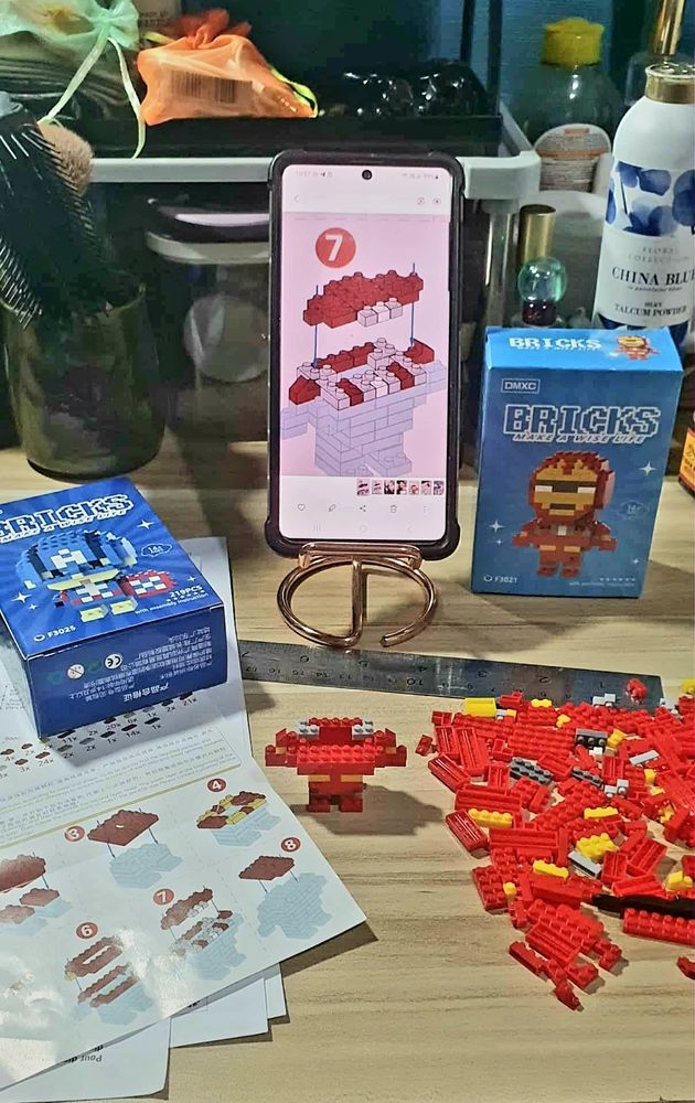 My work desk/dressing table at 11 pm tonight: 

Two small cardboard boxes labeled BRICKS: Make A Wise Life *shrug emoji*, one lying on its back, Lego Captain America on the cover, the other upright, Lego Iron Man in front. 

A pile of tiny red brick pieces scattered on the right in front of the Iron Man box, while the instruction page is pinned by the Captain America box on the left.

In the foreground, a half-finished Iron Man figurine
In the background, on a phone stand, my phone with a zoomed-in screenshot of #6 diagram on the instruction page.  Because combination presbyopia/myopia/photophobia is my curse.