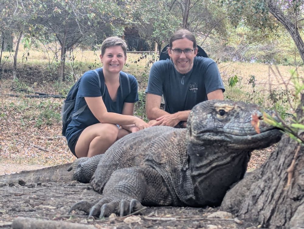 Two extremely attractive people posing behind a large Komodo dragon. The people are wearing grey shirts and smiling. The Komodo dragon looks like it might be smelling a flower, but probably is just resting near the flower. 