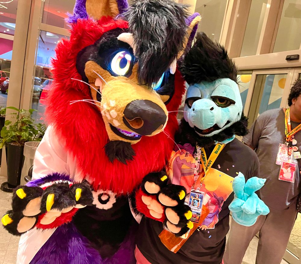 Two cosplayers in furry suits. One is a bulky fuzzy canine-like character with large padded paws, snout, and hairpiece. The eyes on this suit glow with an LCD screen. The other is a smaller reptilian character with lazy eyes and a fluffy beard. He is making a peace sign. 