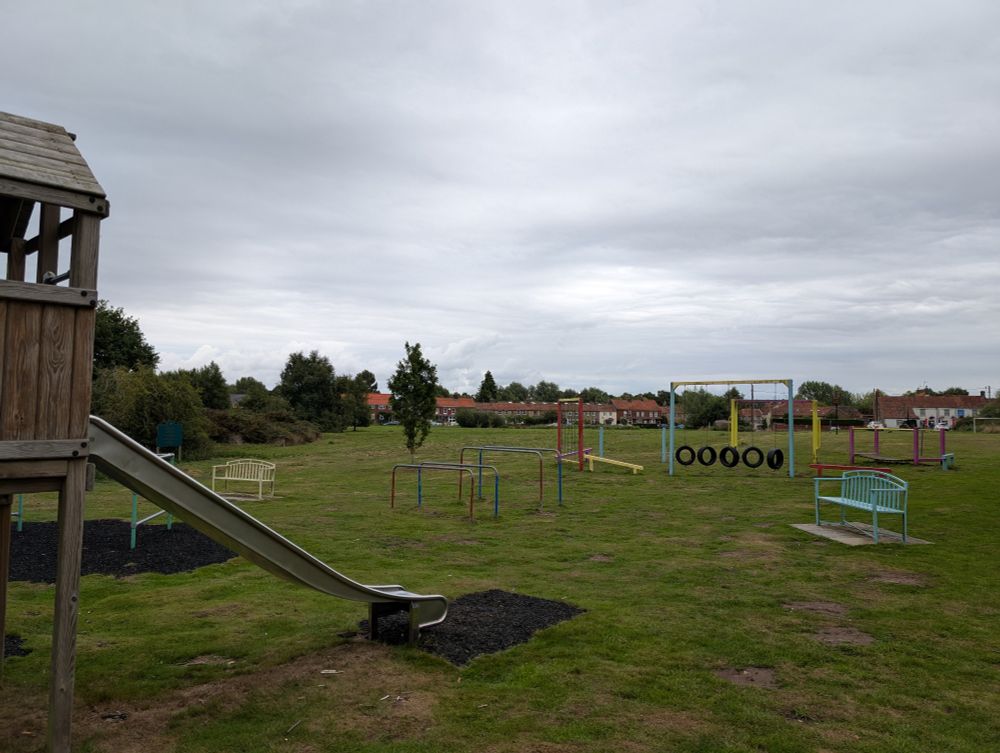 Play equipment, benches in front of common and a row of cottages in the far distance.