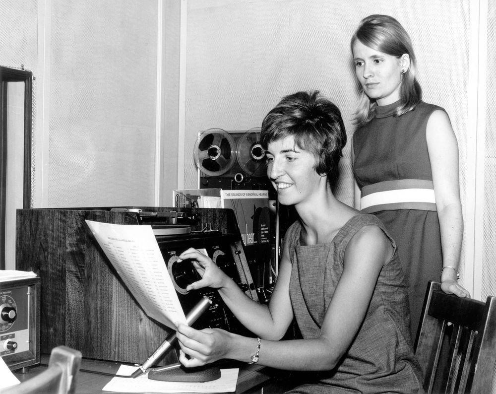 Black and white image of two women with equipment. One is seated, holding a paper and turning a dial, and the other is standing behind her.