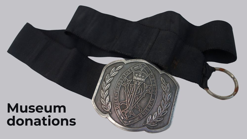 Image of a nurse's belt made of dark blue, ribbed fabric with a large silver buckle featuring the 'VRI' [Victoria Regina Imperatrix] cypher bordered by the title 'Queen's Institute of District Nursing', and accompanied by laurel wreath designs. To the left of the belt is text reading 'Museum donations'.