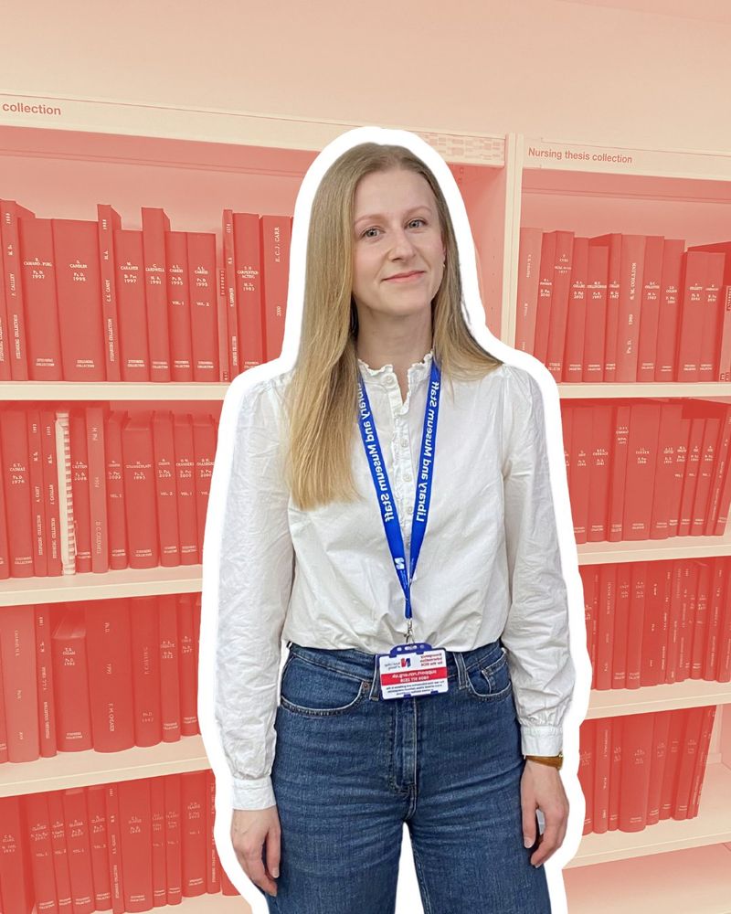 Steph stands in front of a shelf of books labelled "Nursing thesis collection". She is smiling and wearing a white blouse, blue jeans and her 'Library and Museum' lanyard.