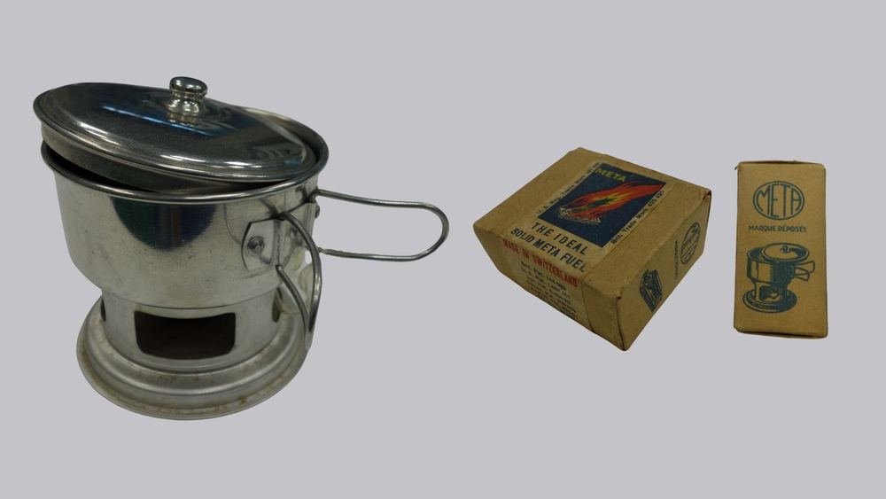 An image featuring a late-20th century Meta brand stove consisting of three silver coloured parts - lid, pot with handles and fuel stand.

To the right of the stove is an image of a little rectangular box, the packaging for 'The Ideal Solid Meta Fuel'. Above the name is a colourful graphic of one of the fuel tablets aflame. The bottom of the box is visible showing the 'made in Switzerland' label and patent numbers for Britain, U.S., India, Canada and two more which are not readable in the image.

Furthest to the right of the image is a side view of the fuel box, showing the brand name 'META' and 'marque déposée' [registered trademark] and an illustration of the stove mentioned above.