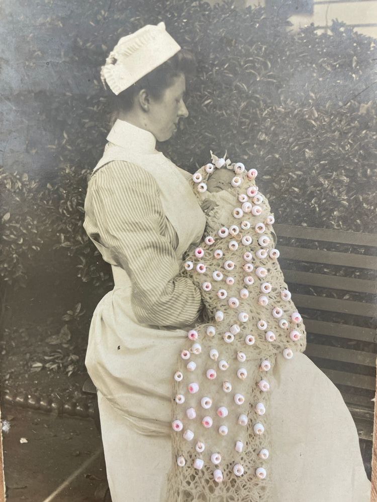 Vintage black and white photograph depicting a nurse sitting on a bench and looking at a baby in her arms, embellished with pink beads.