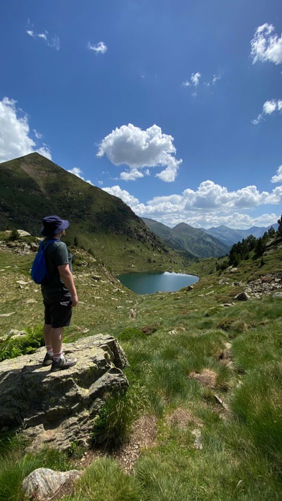 I am standing on a rock overlooking a deep green valley that leads down to a lake. There is a mountain range in the distance. The location is Andorra.