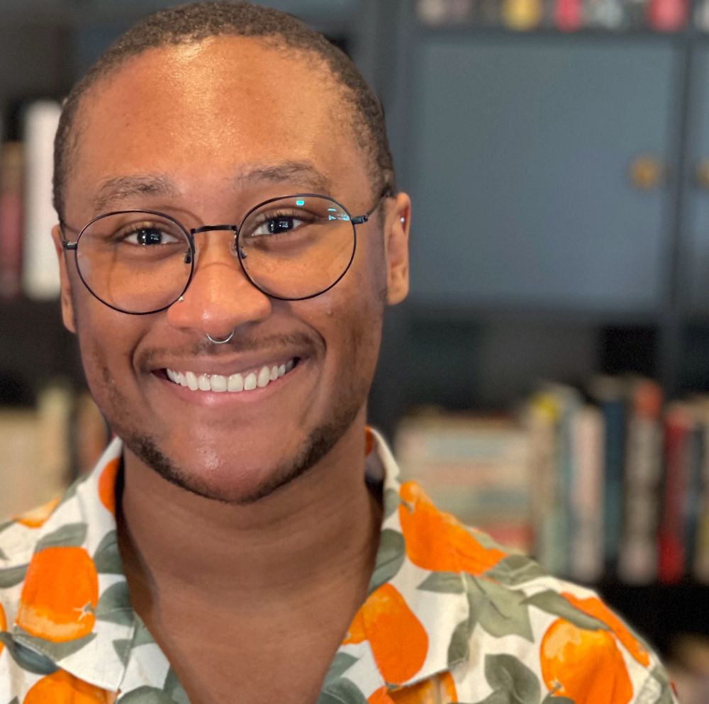 Photo of the author, a smile on their face. They wear a casual shirt with images of oranges. 
