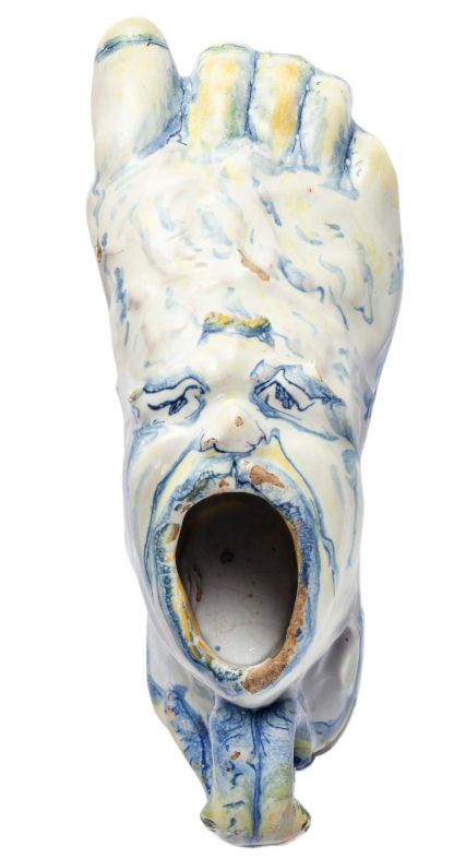 Inkwell in the form of a foot. The mouth of the inkwell is painted to resemble a human mouth, with a human face, or mask, painted on top of the foot.