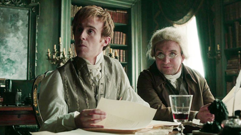 A screen grab of Henry Lascelles and Gilbert Norrell in the miniseries based on the novel "Jonathan Strange & Mr Norrell." They are sitting at a table, looking up from some papers.