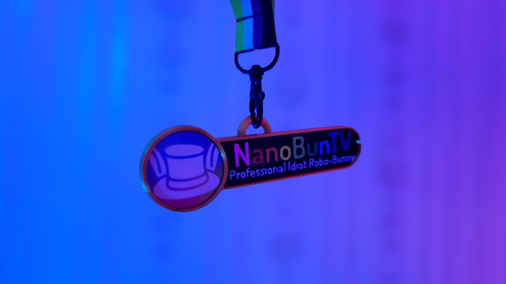 A VRChat styled nametag with the name "NanoBunTV" and a lower subtitle/status text that reads "Professional Idiot Robo-Bunny". The nametag is hanging from a nonbinary lanyard.