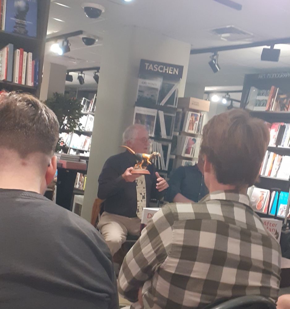 Dougal Dixon in a bookshop holding a model of a Cutlasstooth (Caedosaurus gladiadens)