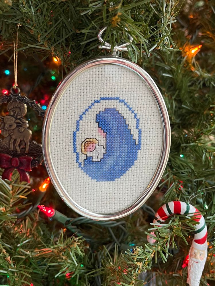A cross-stitched ornament of baby Jesus being enveloped by a large blue object that looks like an ocean wave but that is, on further inspection, the Virgin Mary in a blue cloak
