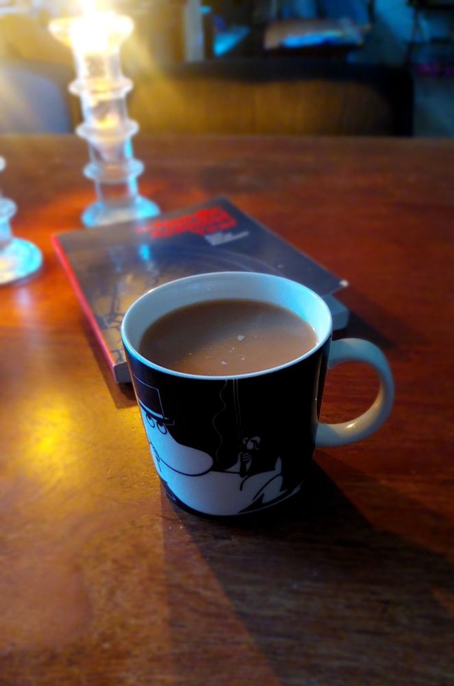 Eldheitur tebolli með hunangi og haframjólk. Bollinn er svartur með mynd af Múmínpabba. Í bakgrunni er kertaljós og bókin Nordic Animist Calendar eftir Rune Rasmussen. 