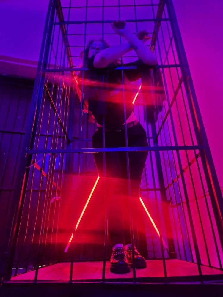 foto tirada do chão para cima. estou em uma gaiola de metal, em pé, com os braços pra fora da grade. visto uma calça preta e blusa preta com mangas largas na ponta. atrás de mim tem uma luz de led vermelha em formato de X