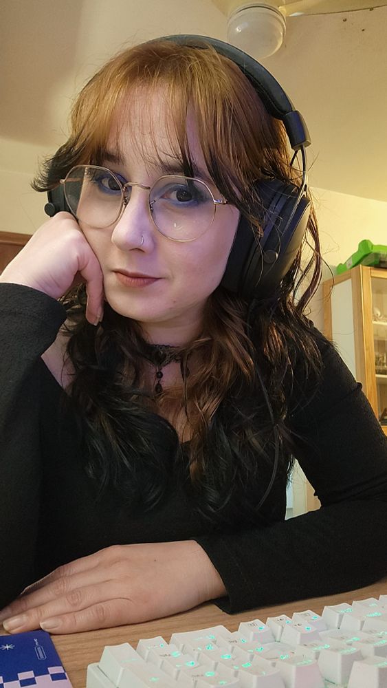 selfie minha, de óculos, encostada na escrivaninha, com fones de ouvido e com a mão no rosto.