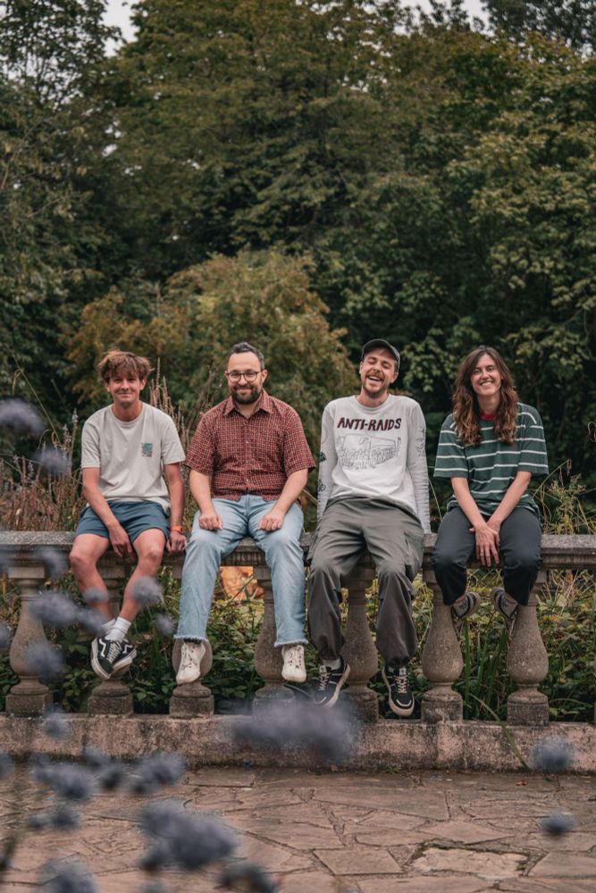 the four members of adults sat laughing and smiling on a wall (kinda? it's got little pillars holding up a long strip of stone, not sure what they're called) in a flower garden at a park. trees behind them, stone floor in front.