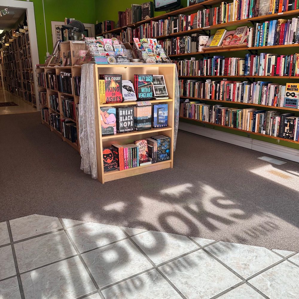 interior photo of a bookstore, we see light wood bookshelves and a shadow on the floor that says BOOKS AND MORE, NEW AND USED