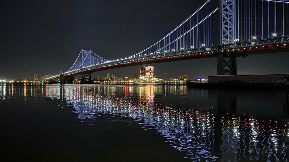A picture of the Benjamin Franklin Bridge over the Delaware River at night in Philadelphia, Pennsylvania. Lit up buildings in New Jersey are visible in the distance. The bridge is lit up and the lights of the bridge and buildings reflect in the water. The water is relatively calm. The sky is a dark gray due to the light pollution of the city.